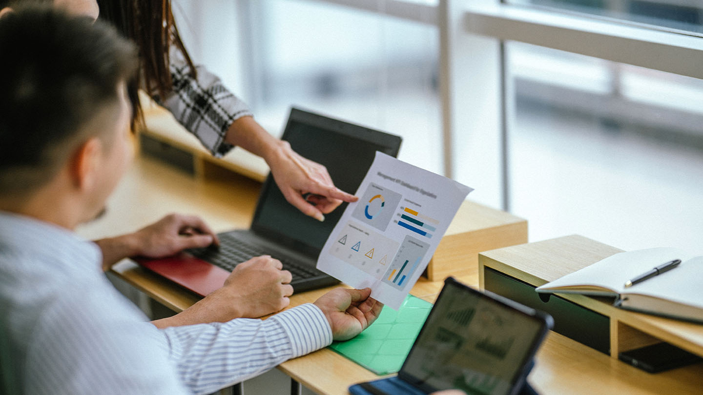 Two people reviewing a printed business report with charts and graphs at a desk with laptops.